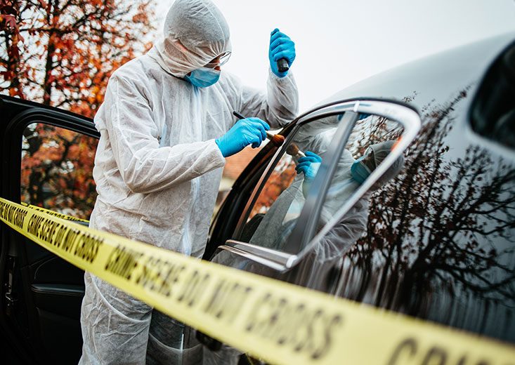 forensics technician wiping for prints on car behind crime scene tape